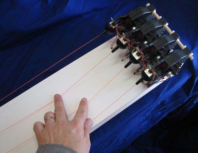 A hand presses four red threads that run along a long white board from four Gametrak tether sensors mounted in a row at its far end.